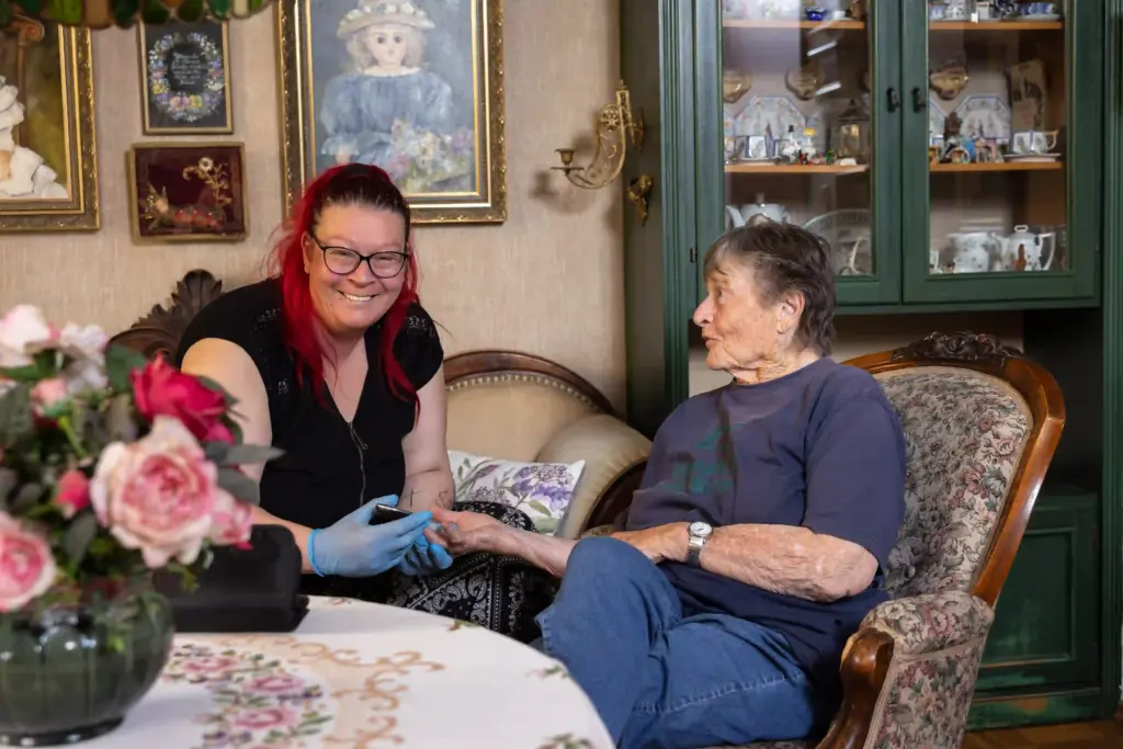 Pflegerin mit Einmalhandschuhen betreut eine Seniorin im Wohnzimmer und hält ihre Hand